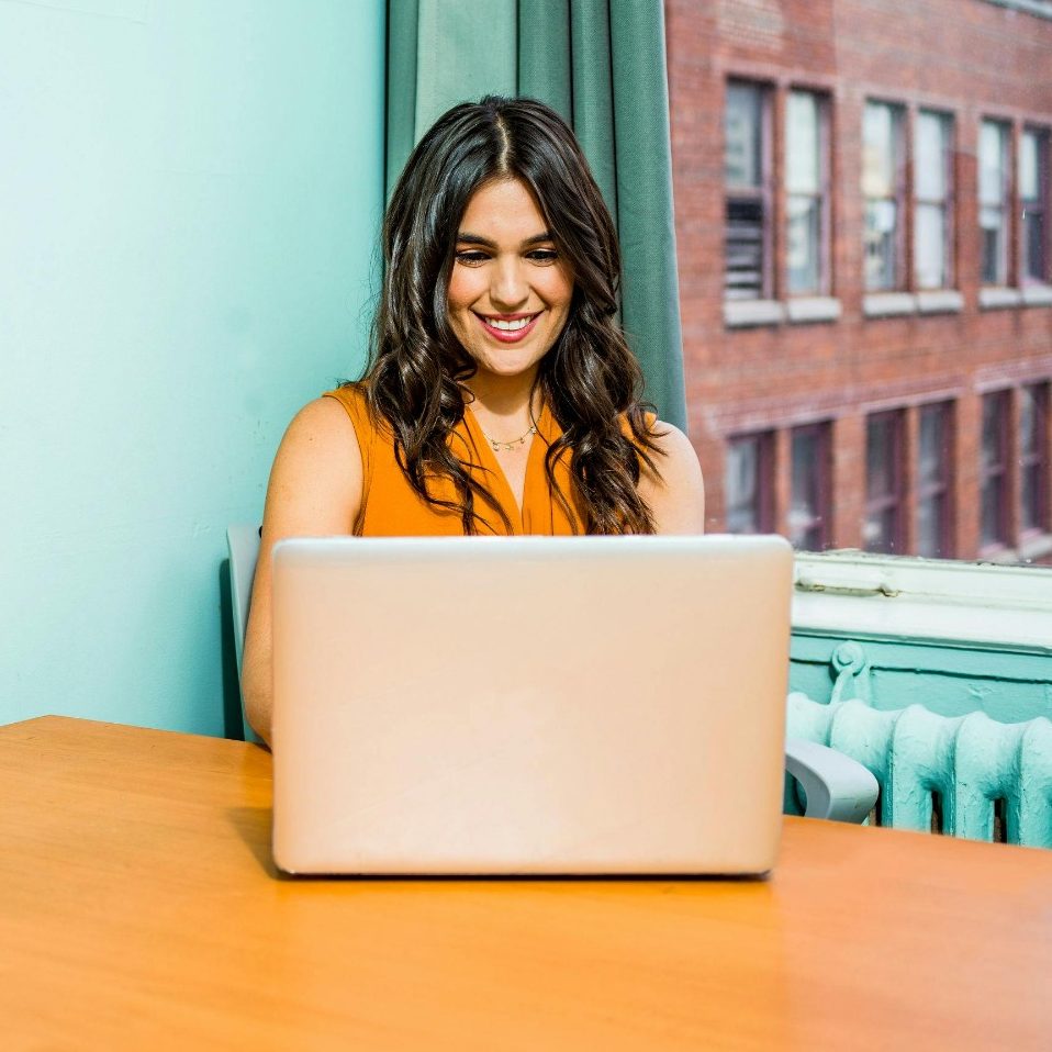 Frau in orangefarbener Bluse sitzt an einem Tisch und arbeitet am Laptop.
