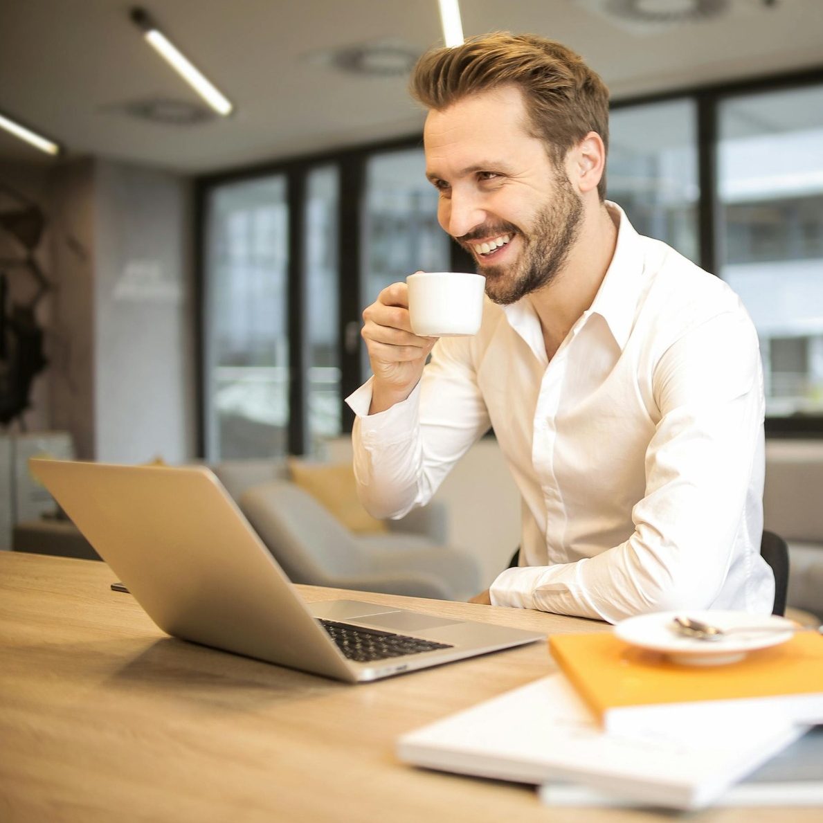 Mann mit Kaffeetasse sitzt lächelnd vor einem Laptop in modernem Büro.