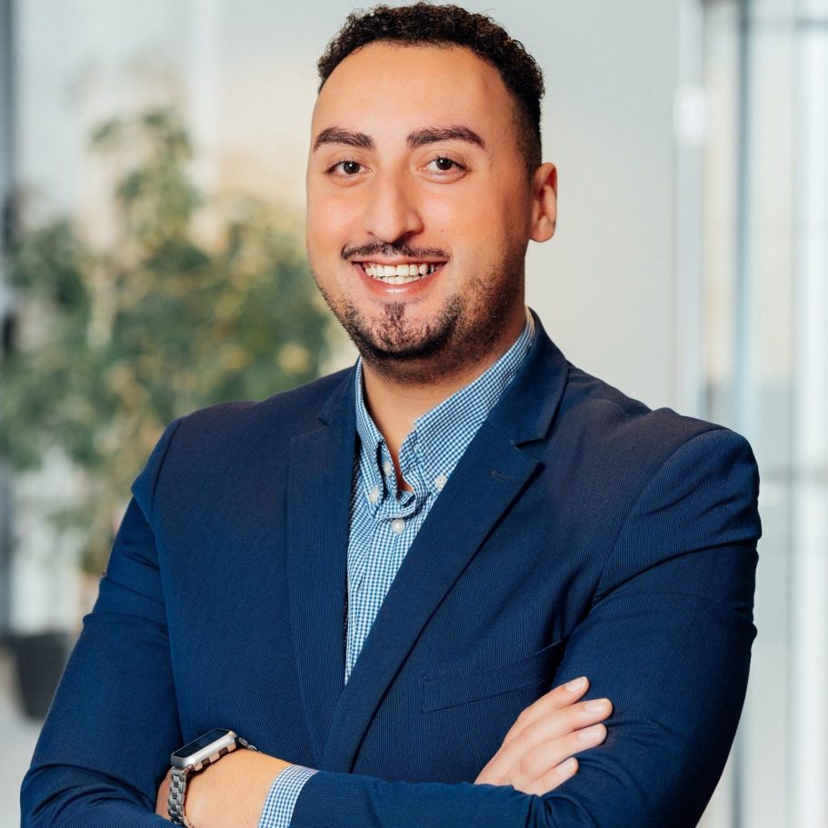 Mann mit dunklem Haar, lächelnd, trägt einen Anzug und steht in einem modernen Büro.