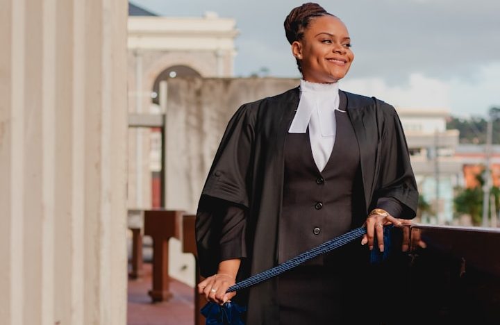 Junge Anwältin in Roben, lächelnd und selbstbewusst, vor historischem Hintergrund. BU für Kammerberufe