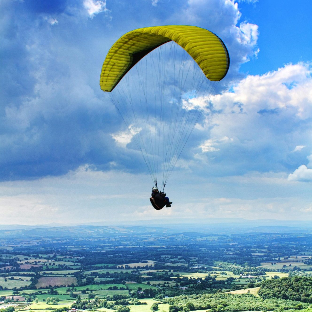 Person mit gelbem Gleitschirm schwebt über einer grünen Landschaft unter blauen Himmel.
