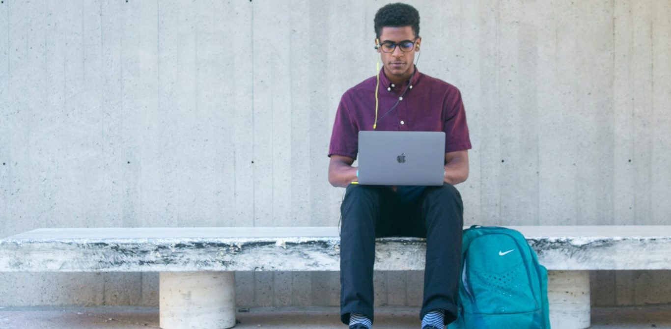 BU für Auszubildende & Azubis Junger Mann mit Laptop sitzt auf einer Bank, im Hintergrund eine graue Wand. BU für Auszubildende & Azubis