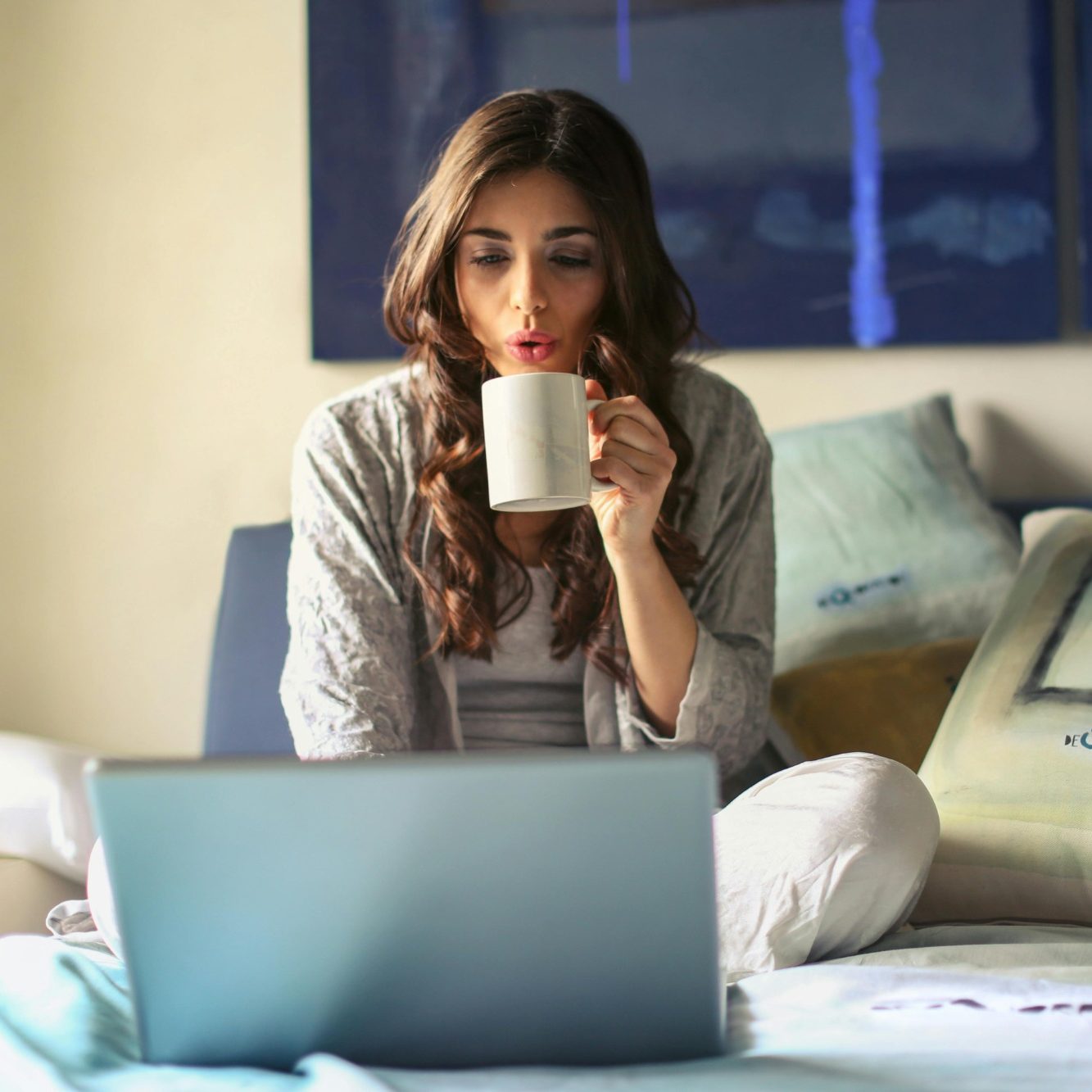Frau mit langen Haaren sitzt auf dem Bett und trinkt aus einer Tasse, während sie auf einen Laptop schaut.