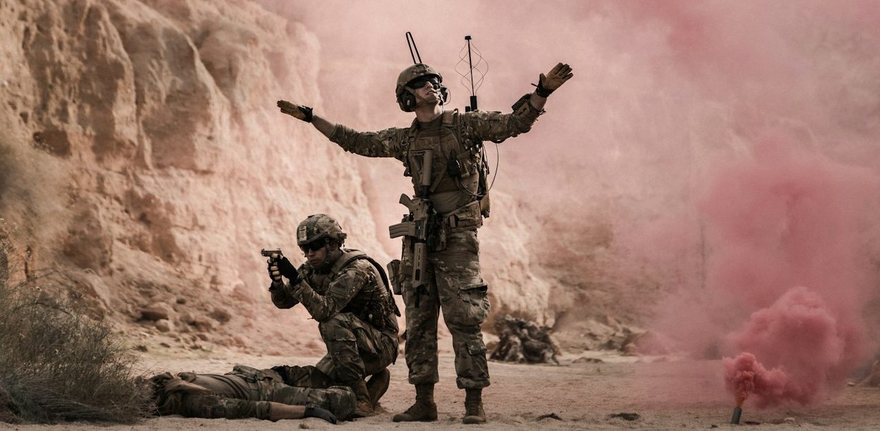 Soldaten in unruhiger Umgebung, einer im Vordergrund mit erhobenen Armen, rosa Rauch. BU für Soldaten