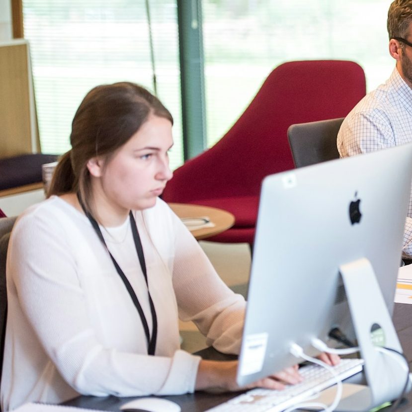 BU für Kaufleute & Büroberufe Eine Frau arbeitet konzentriert am Computer, während ein Mann im Hintergrund sitzt zur Berufsunfähigkeitsversicherung