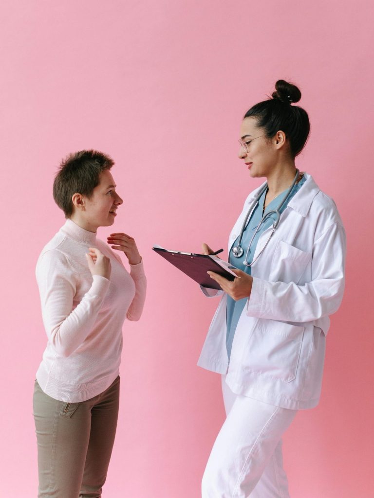 Ärztin spricht mit Patientin, beide stehen vor pinkem Hintergrund.