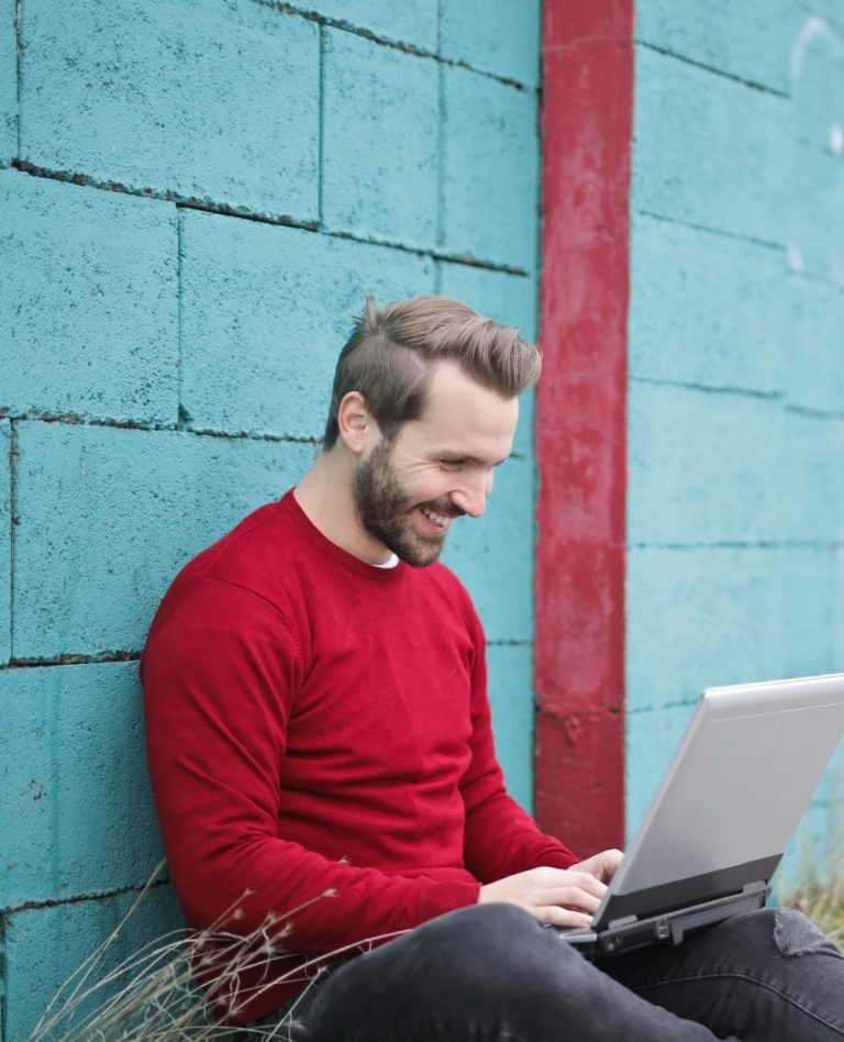 Mann in rotem Pullover sitzt an einer Wand und arbeitet lächelnd am Laptop.