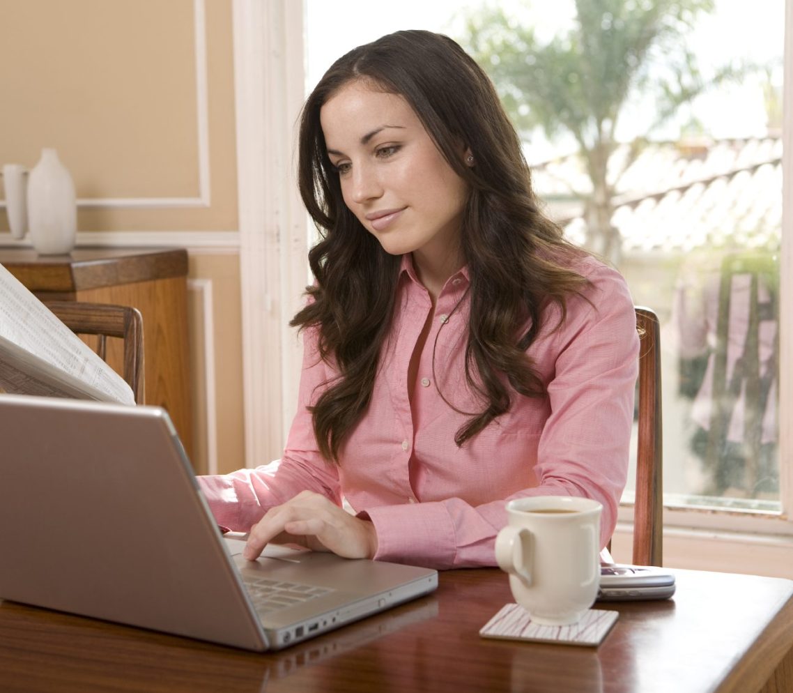 Eine Frau in einem rosa Oberteil sitzt an einem Tisch und arbeitet am Laptop.