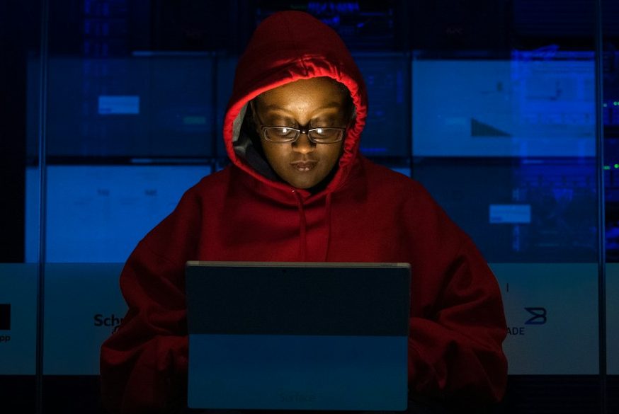 BU für IT-Berufe Eine Person in rotem Hoodie sitzt vor einem Laptop, umgeben von dunklem, futuristischem Hintergrund. BU für IT-Berufe