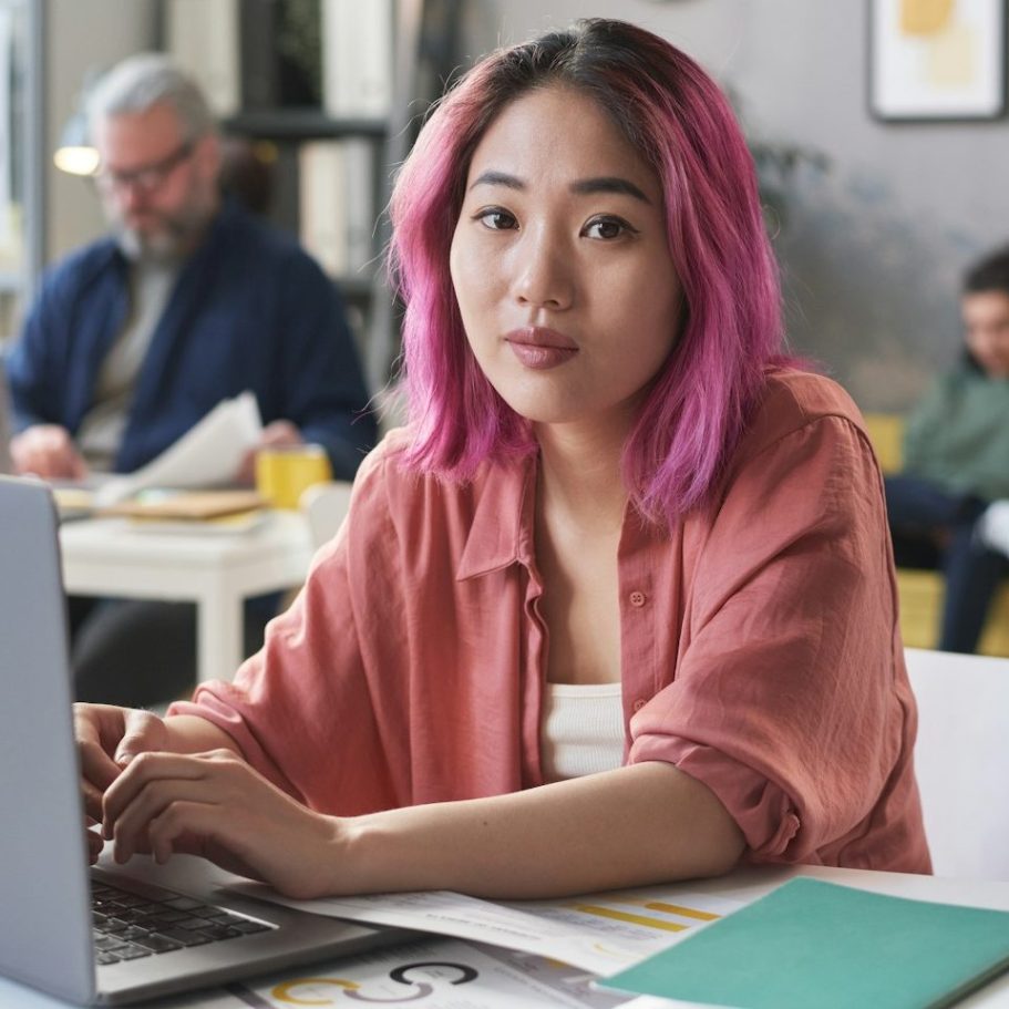 BU für Studenten Junge Frau mit pinken Haaren arbeitet am Laptop, umgeben von Personen in einem Café zur Berufsunfähigkeitsversicherung