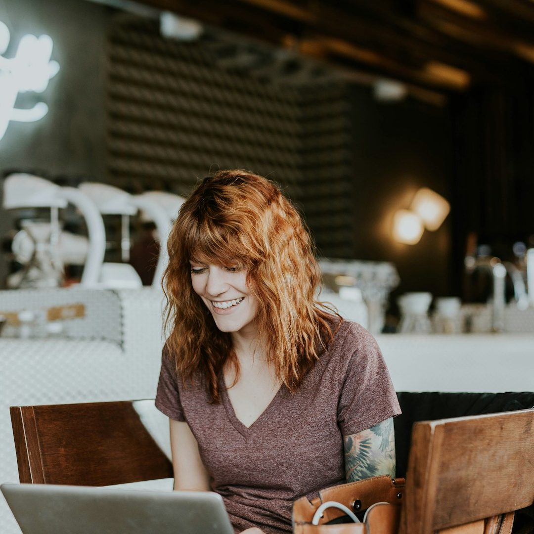 Berufsunfähigkeitsversicherung Blog Frau mit lockigem, rotem Haar sitzt lächelnd am Laptop in einem Café und schaut sich den BU-Direkt Blog für Berufsunfähigkeitsversicherung