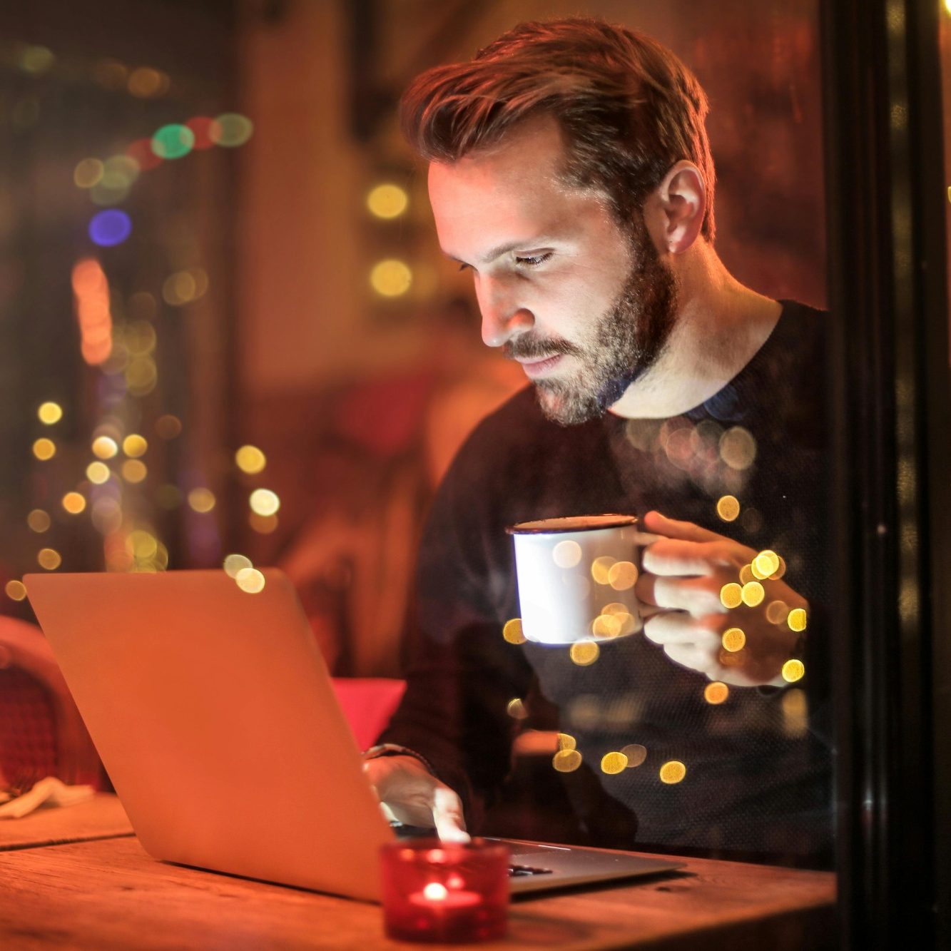 Mann am Laptop, während er eine Tasse hält, umgeben von gemütlicher Beleuchtung.