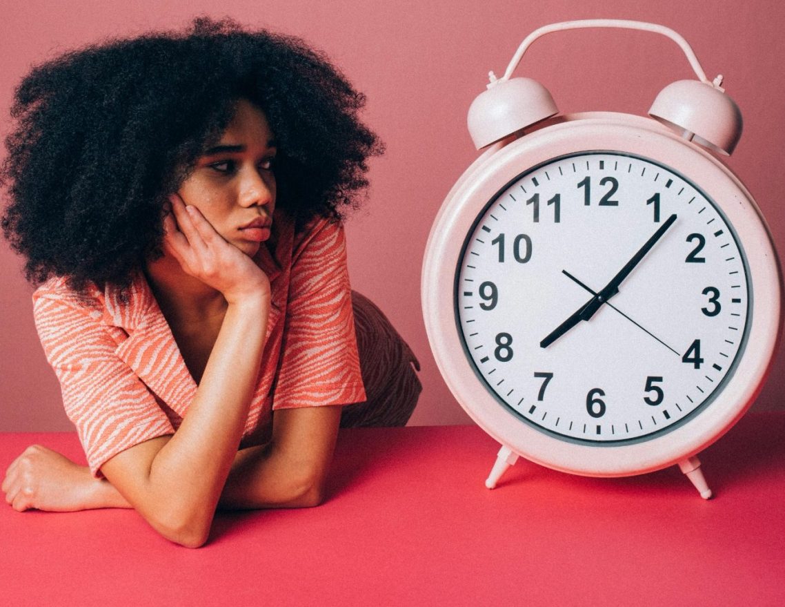 Junge Frau mit lockigem Haar, nachdenklich neben einer rosa Wanduhr.