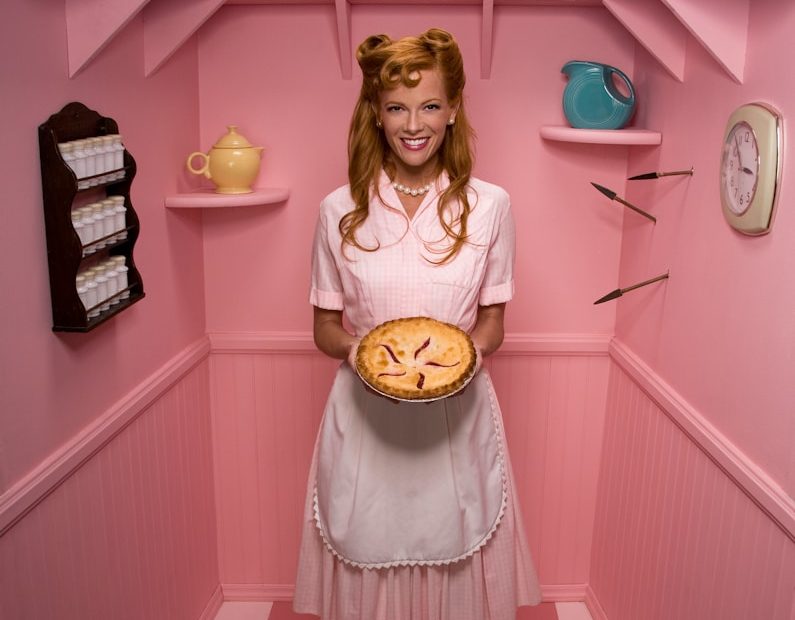 U für Hausfrauen und Hausmänner Frau in rosa Schürze hält eine Kuchenschale in einer pinken Umgebung. BU für Hausfrauen und Hausmänner