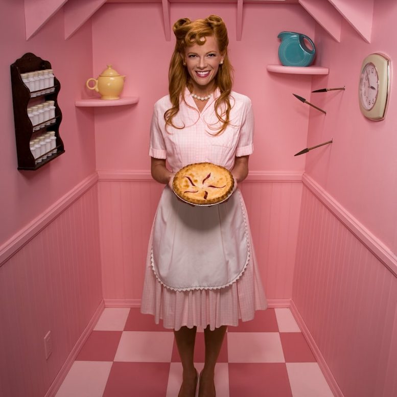 BU für Hausfrauen & Hausmänner Frau in rosa Kleid hält einen Kuchen in einer pastellfarbenen, nostalgischen Küche zur Berufsunfähigkeitsversicherung