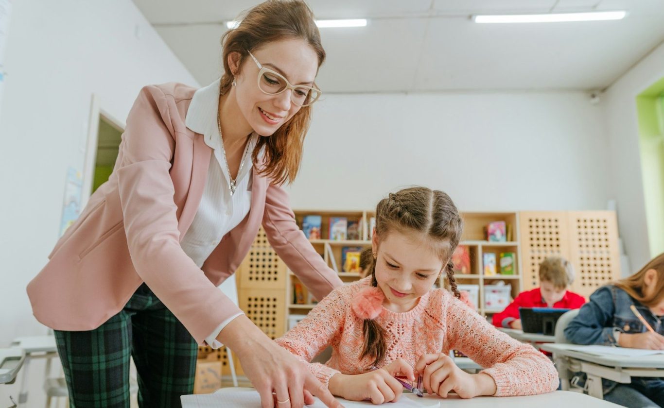 BU für Lehrer und Referendare Lehrerin hilft einem Mädchen beim Schreiben in einem Klassenraum. Kinder sind im Hintergrund. BU für Lehrer und Referendare