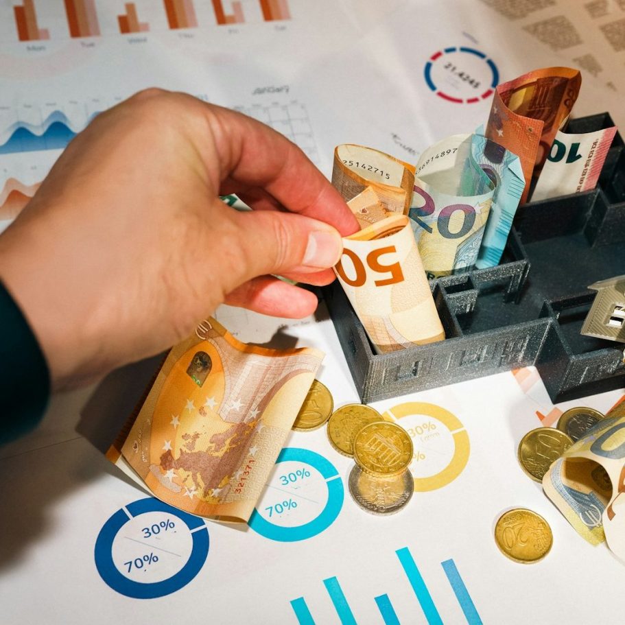 Hand legt Geldscheine in eine Kasse, umgeben von Münzen und Diagrammen.