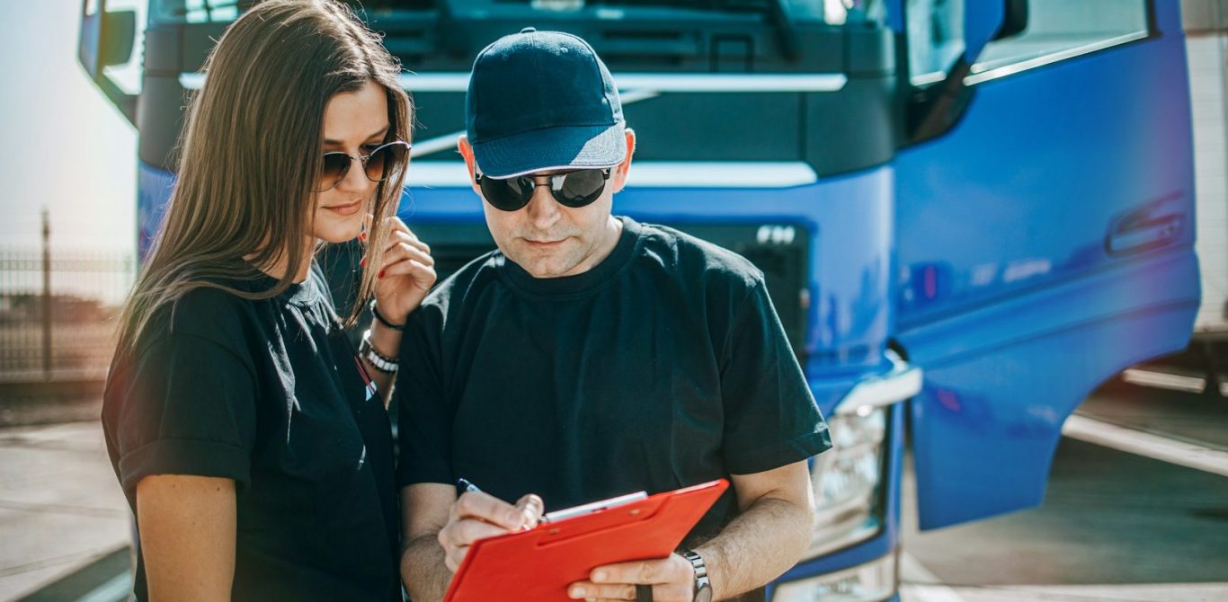 Zwei Personen, eine Frau mit Sonnenbrille und ein Mann, überprüfen Dokumente vor einem Lkw. BU für LKW und Busfahrer
