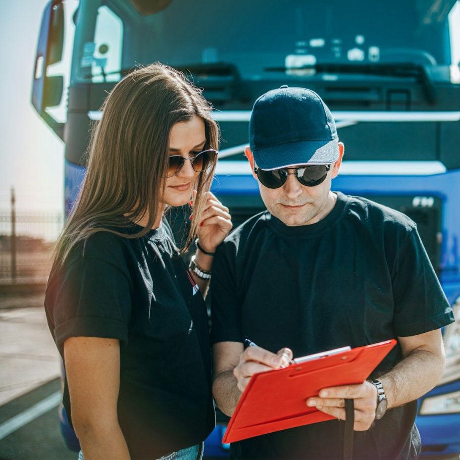 BU für LKW & Busfahrer Zwei Personen in T-Shirts, einer schreibt auf ein rotes Clipboard vor einem Lkw zur Berufsunfähigkeitsversicherung