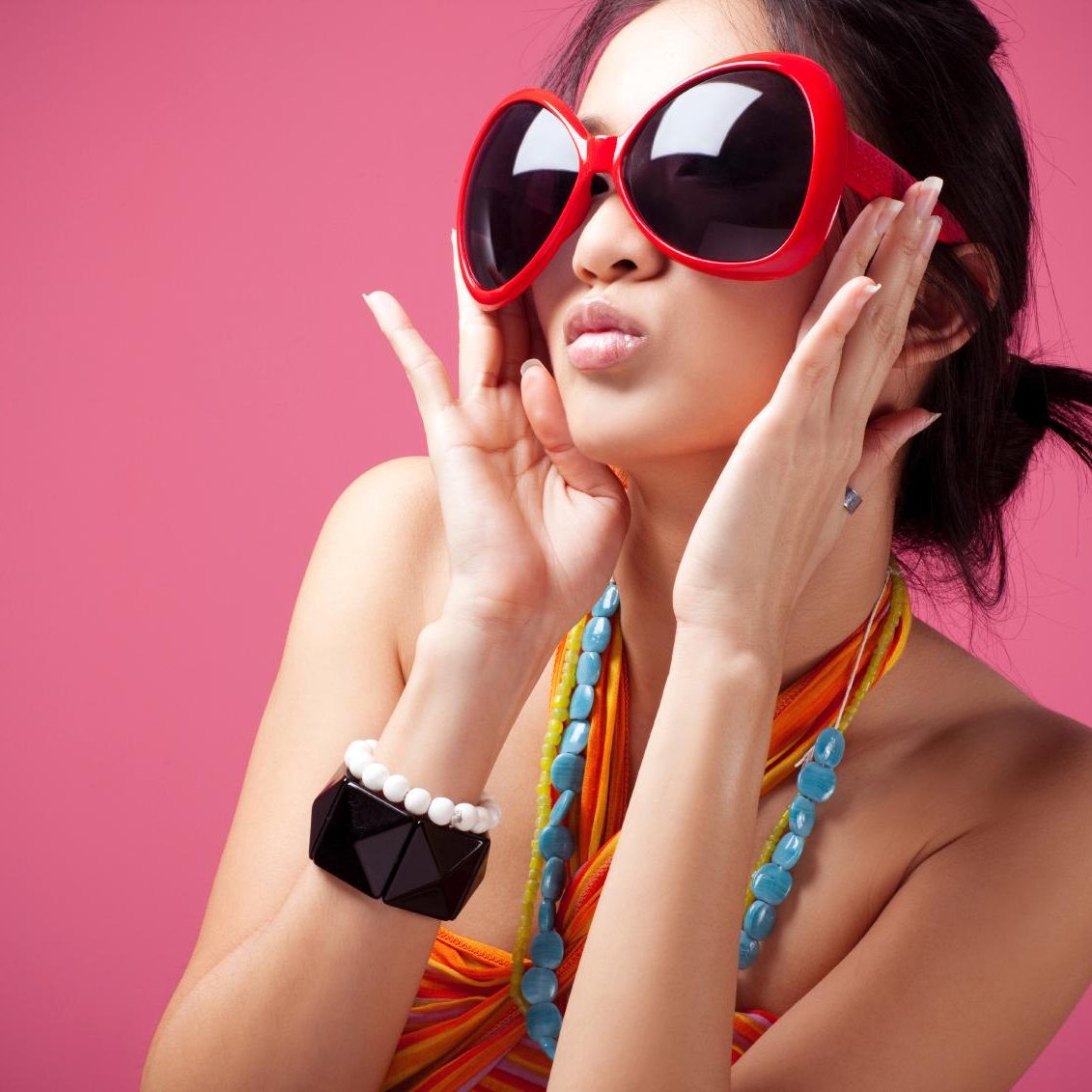 Frau mit großen, roten Sonnenbrillen und bunten Accessoires vor pinkem Hintergrund.