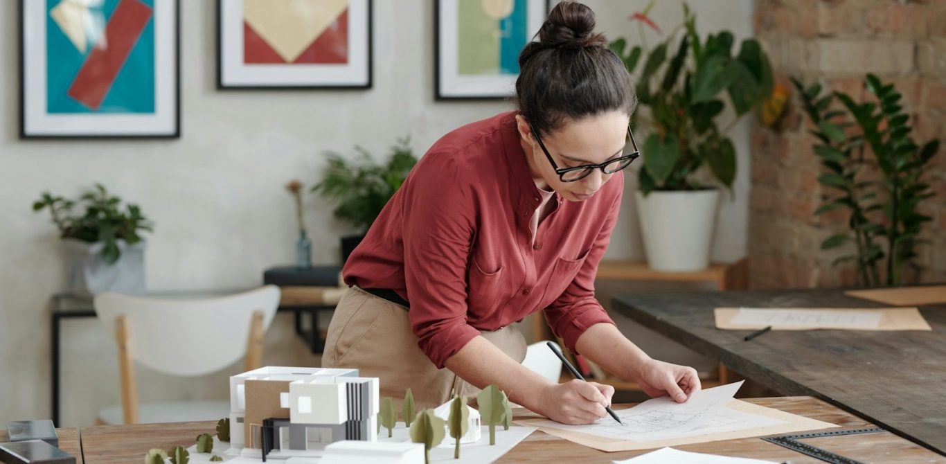 Frau in Brille zeichnet Skizzen an einem Tisch mit Pflanzen und Kunstwerken im Hintergrund.  BU für Ingenieure