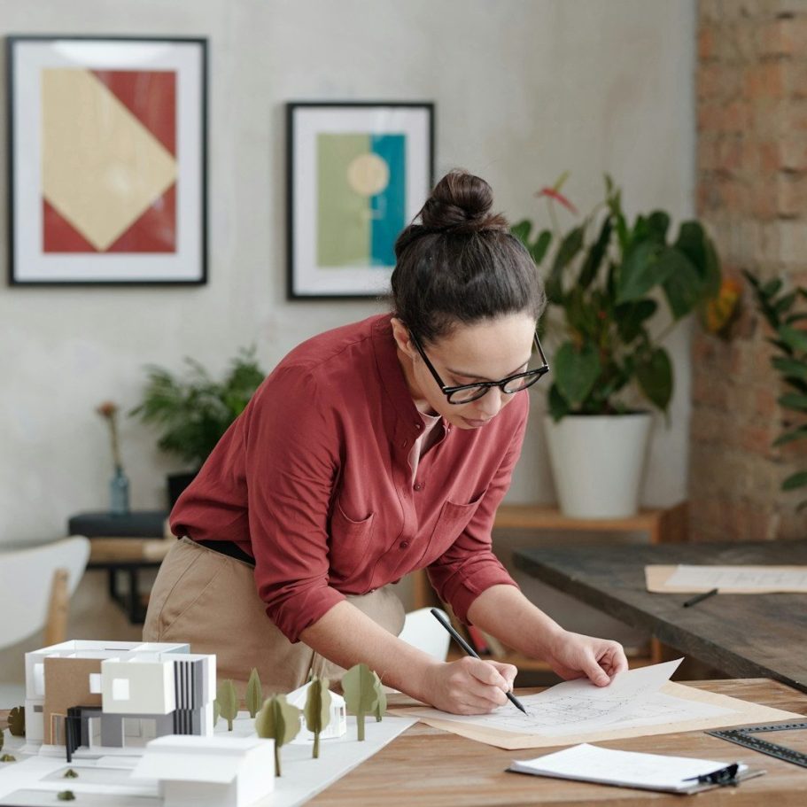 BU für Ingenieure Frau mit Brille, die an einem Tisch an einem Designprojekt arbeitet. Pflanzen im Hintergrund zur Berufsunfähigkeitsversicherung