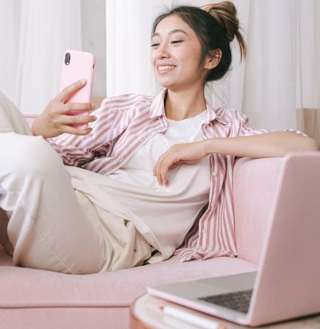 Junge Frau mit Smartphone, entspannt auf einer pinken Couch, neben einem Laptop. Online-Beratung zur Berufsunfähigkeitsversicherung