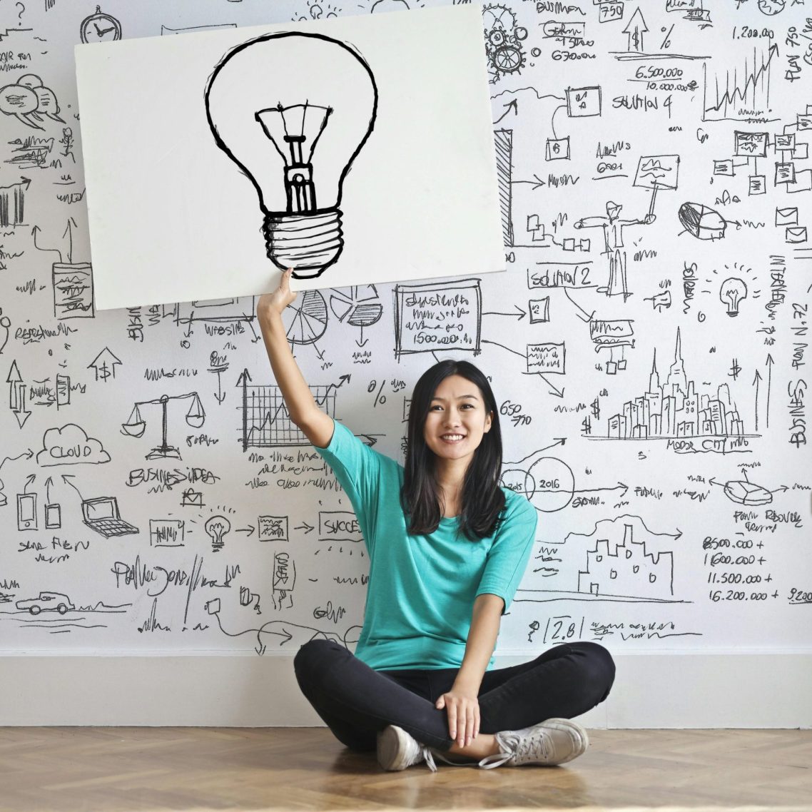 Frau mit einem Leuchtmittel-Symbol, sitzt vor einer kreativ gestalteten Wand.